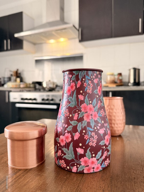 Decorate water jar made from copper with red and pink floral design sitting on a brown surface with two copper cups in the background