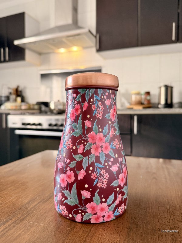 Decorate water jar made from copper with red and pink floral design sitting on a brown surface in a kitchen