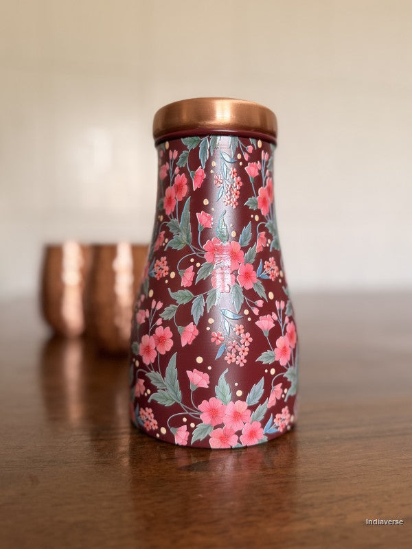 Decorate water jar made from copper with red and pink floral design sitting on a brown surface with two copper cups in the background