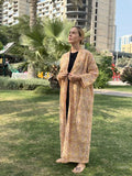 Person wearing a long, patterned robe standing in a park with buildings and palm trees in the background.