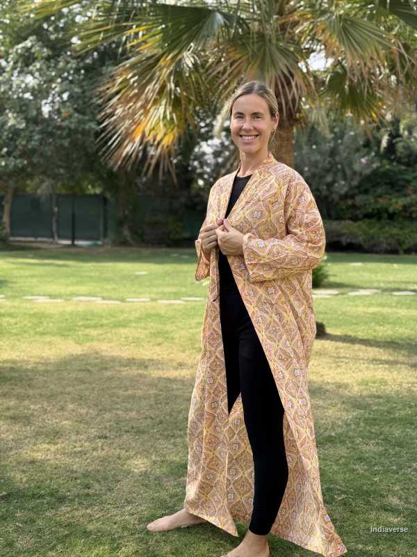 Woman wearing a long, patterned beige cardigan over a black outfit in an outdoor setting with greenery.