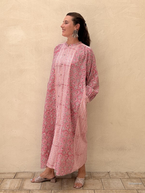 A woman wearing a long-sleeved pink and red floral print kaftan dress, standing against a textured beige stucco wall.