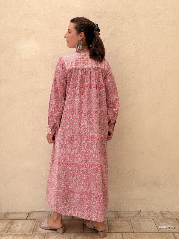 A woman wearing a pink and white patterned long-sleeve maxi dress, shown from the back against a textured beige stucco wall.