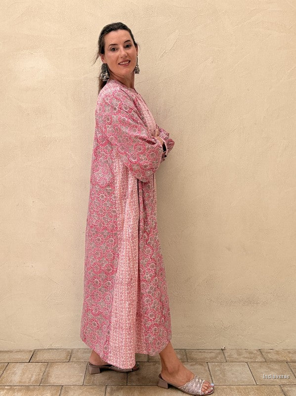 A woman wearing a long pink patterned kaftan dress with a tie waist, standing against a textured beige stucco wall.