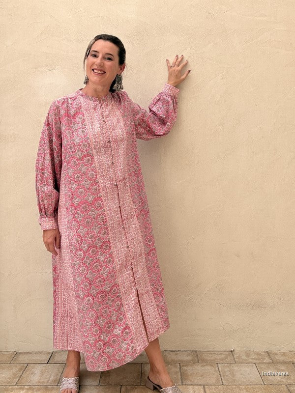A woman wearing a pink and white patterned long-sleeve tunic dress stands against a textured beige stucco wall, holding her hand on the wall.
