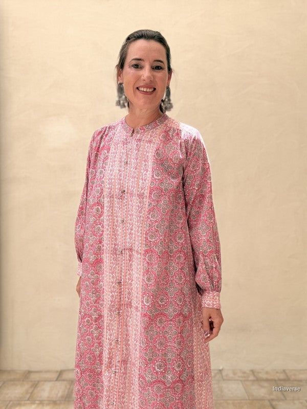 A woman wearing a pink and white patterned long-sleeve tunic dress, standing against a beige textured wall.