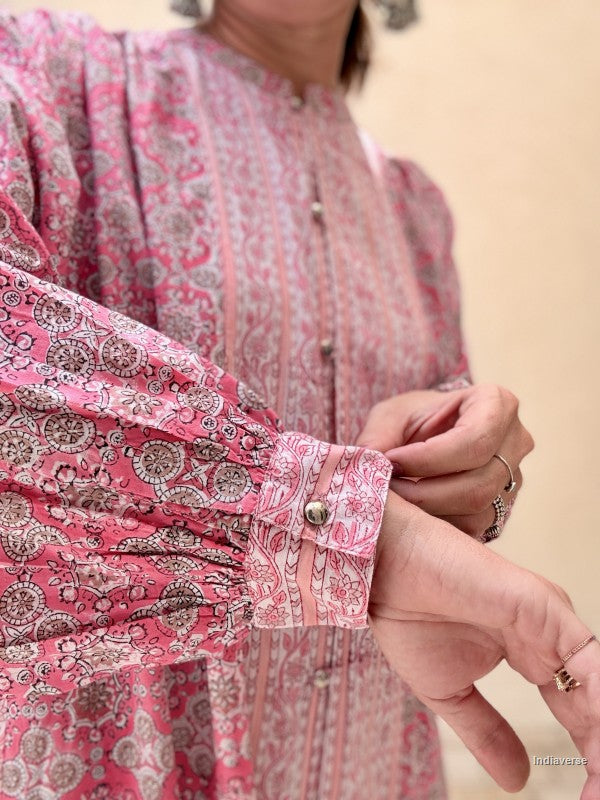 A close-up of a person wearing a pink and beige paisley-patterned blouse, adjusting the cuff of the sleeve. The blouse features a high neckline, vertical button placket, and gathered sleeves.