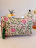 A quilted cosmetic pouch with a pink and green floral vine pattern on a beige background, featuring a green tassel zipper pull, set against a softly blurred background with bathroom or vanity items | Rose Garden Beige