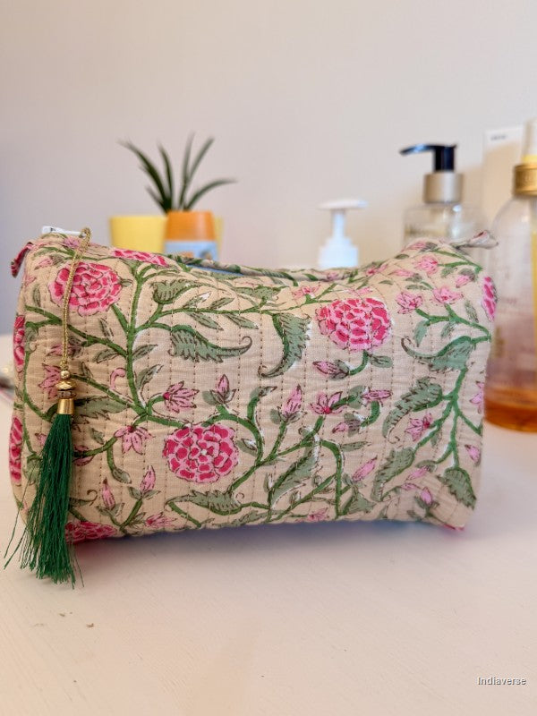 A quilted cosmetic pouch with a pink and green floral vine pattern on a beige background, featuring a green tassel zipper pull, set against a softly blurred background with bathroom or vanity items | Rose Garden Beige