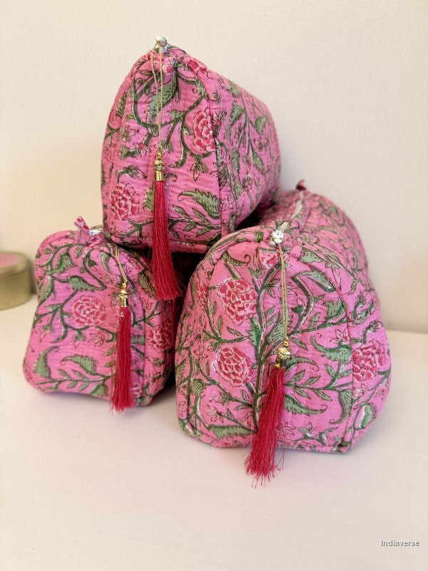 Three small drawstring cosmetic pouches made of pink floral batik fabric with green leaves, red tassels, and gold-toned zipper pulls, arranged on a light surface against a plain light background | Rose Garden