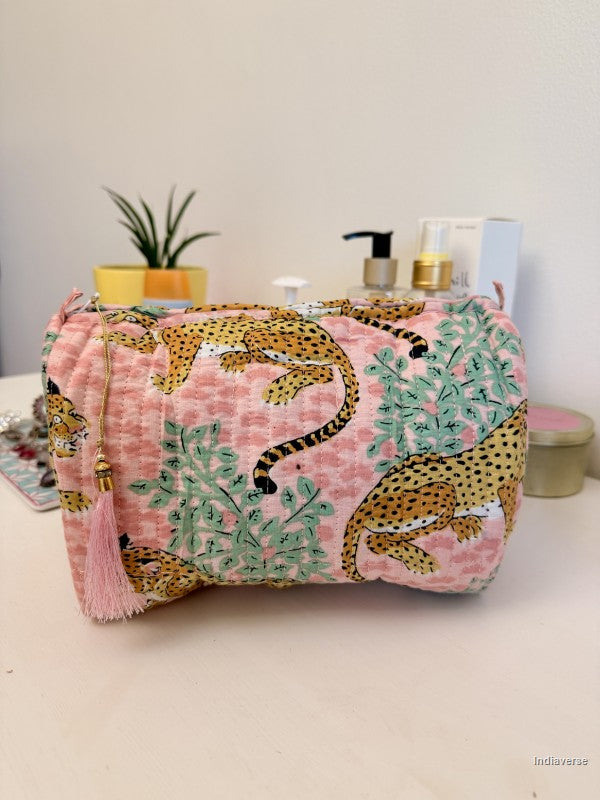 A pink cosmetic pouch with a leopard and green foliage print, featuring a pink tassel zipper pull, displayed on a tabletop with toiletries in the background | Rose Leopard
