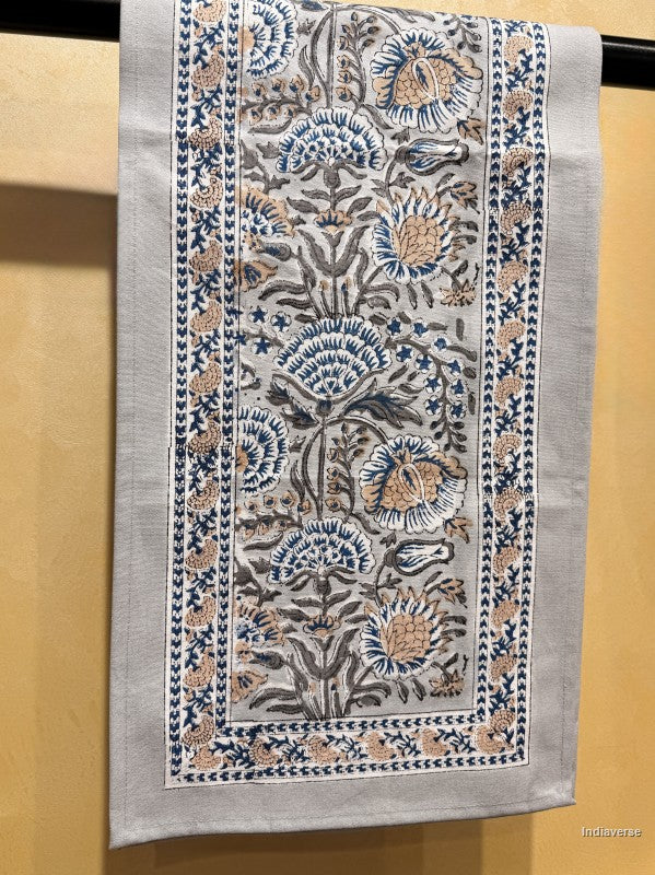 A rectangular embroidered table runner with a floral pattern in blue, beige, and gray on a light gray base, hanging against a warm beige wall.