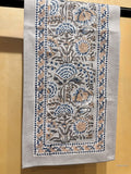 A rectangular embroidered table runner with a floral pattern in blue, beige, and gray on a light gray base, hanging against a warm beige wall.