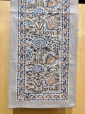 A rectangular handloom textile runner with intricate floral embroidery in blue, beige, and brown on a light gray base, displayed flat against a warm yellow background.