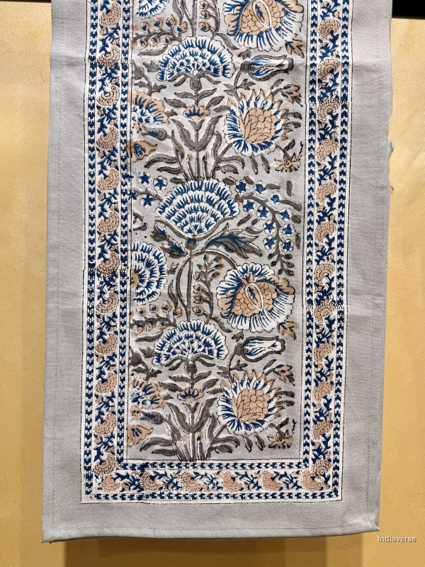 A rectangular handloom textile runner with intricate floral embroidery in blue, beige, and brown on a light gray base, displayed flat against a warm yellow background.