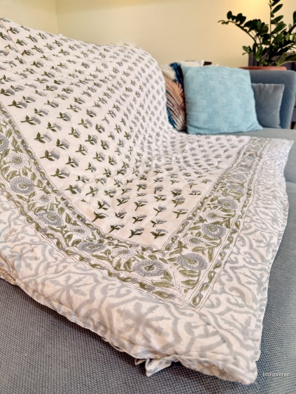 quilted blanket folded on a grey teal colored couch showing a grey geometric pattern on the border and reverse side and on the front small blue and green flowers