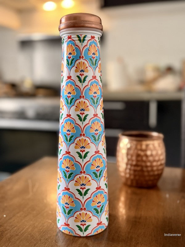 Tall copper water bottle with symmetric floral design in blue and yellow color sitting on a brown table in a kitchen with a copper cup in the background