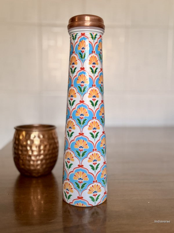 Tall copper water bottle with symmetric floral design in blue and yellow color sitting on a brown table with a copper cup in the background