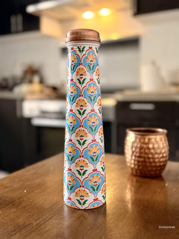 Tall copper water bottle with symmetric floral design in blue and yellow color sitting on a brown table in a kitchen with a copper cup in the background