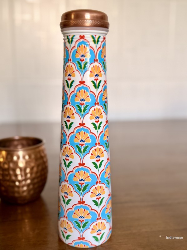 Tall copper water bottle with symmetric floral design in blue and yellow color sitting on a brown table with a copper cup in the background