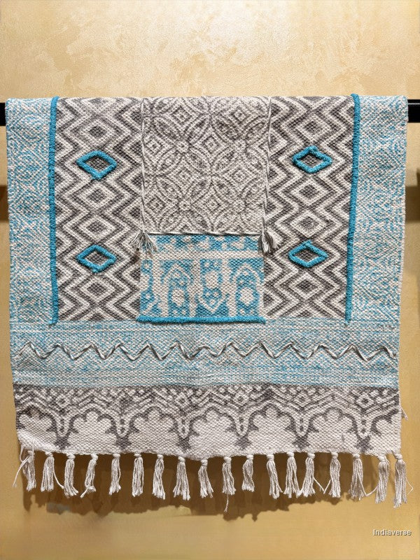 A woven textile with geometric patterns in gray, white, and turquoise blue, hanging flat against a textured beige wall. The textile features a central pocket-like section, diamond motifs, and fringe tassels along the bottom edge.