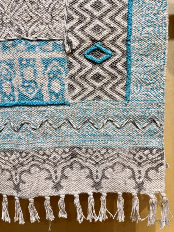 A close-up of a woven textile with geometric and tribal-inspired patterns in gray, white, and turquoise blue, finished with white tassels along the bottom edge.