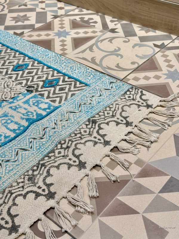 A close-up of a patterned area rug with cream, gray, and turquoise blue geometric and floral motifs, laid over a tiled floor.