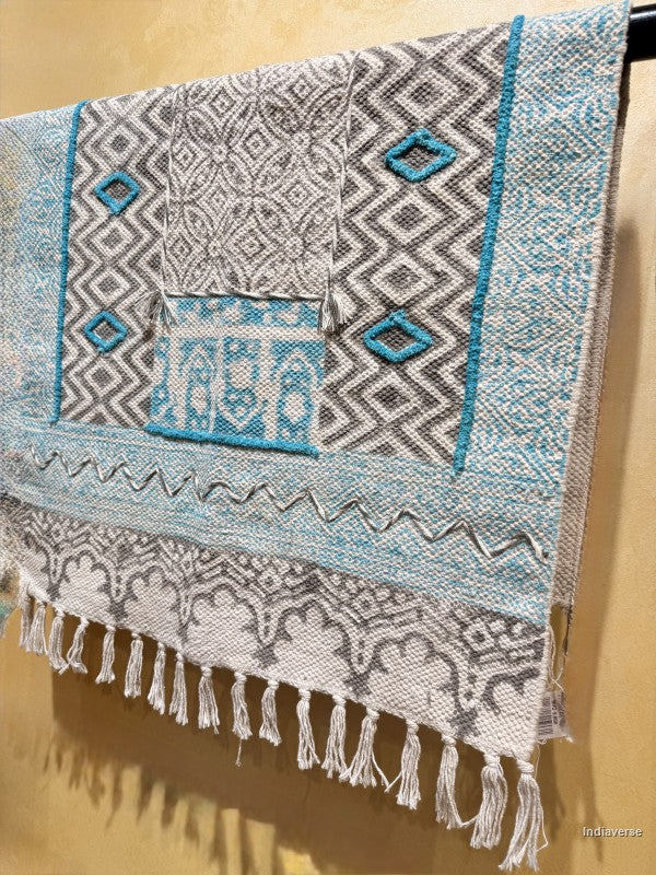 A woven textile with geometric patterns in gray, white, and turquoise blue, featuring a fringed tassel edge, displayed against a warm beige wall.