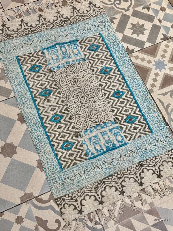 A rectangular patterned prayer rug with fringe, laid diagonally on a tiled floor. The rug features a geometric black and white diamond pattern with turquoise blue borders and accents.