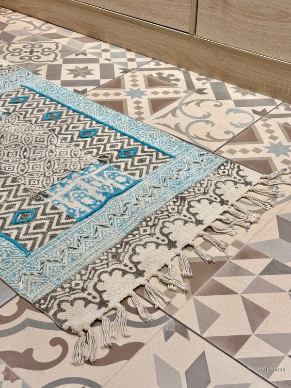 A rectangular woven rug with fringe tassels, featuring a geometric and tribal pattern in cream, gray, black, and turquoise blue, laid out on a patterned tile floor next to a wooden cabinet.