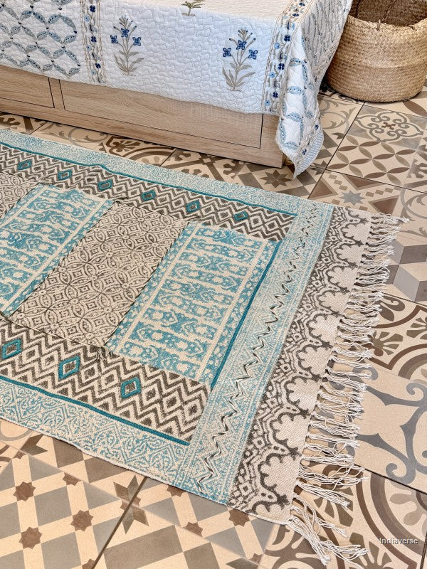A rectangular area rug with intricate geometric and floral patterns in beige, gray, and turquoise blue, styled on a patterned tile floor next to a wooden bed frame.