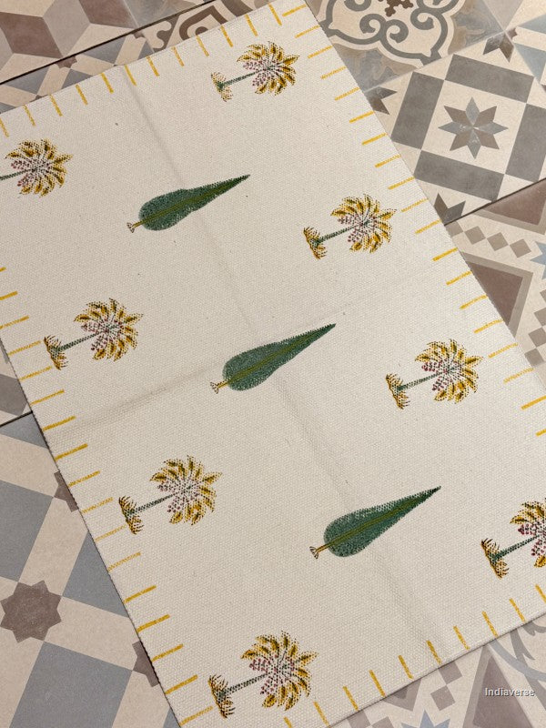 An off-white woven placemat with a printed pattern of green pinecones and yellow-and-white flowers, laid diagonally on a patterned gray and beige tile floor.