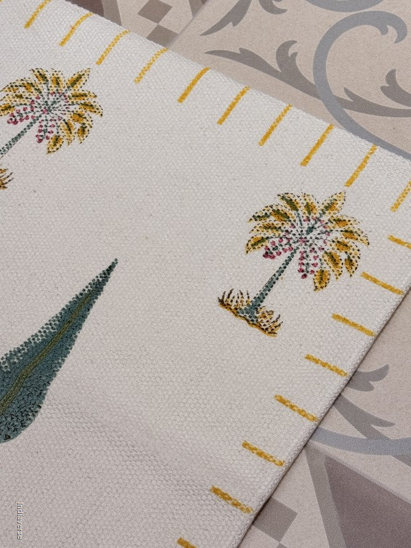 A close-up of a woven textile featuring embroidered palm tree motifs in yellow, green, and pink on an off-white background, with a diagonal fold revealing a patterned gray and beige surface underneath.