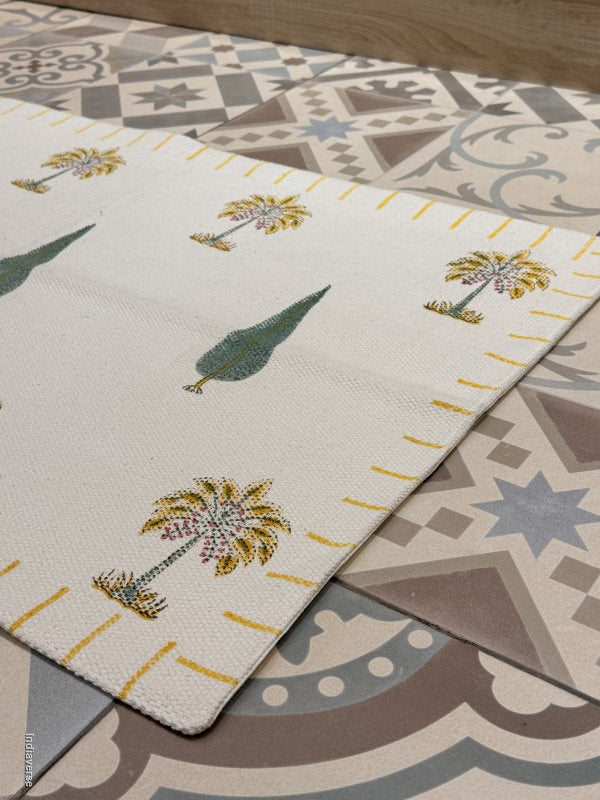 A cream-colored woven runner rug with a pattern of palm trees and green leaves, laid diagonally over a decorative geometric tile floor.