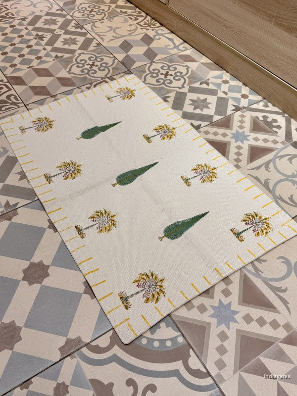 A rectangular cream-colored runner rug with a yellow border and green and yellow palm tree motifs, laid diagonally on a patterned tile floor.