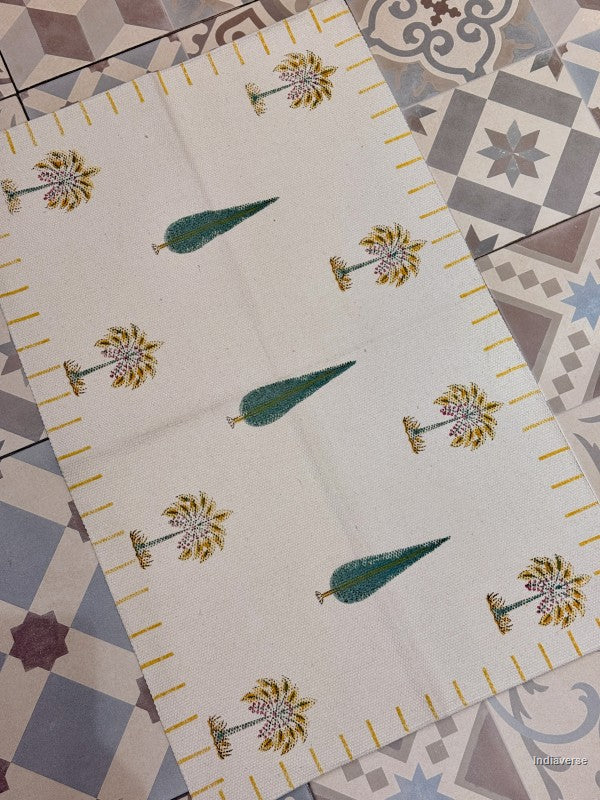 A rectangular cream-colored fabric placemat or runner featuring a repeating block-print pattern of green pine trees and yellow-gold palm trees, laid diagonally on a patterned gray and white tile floor.