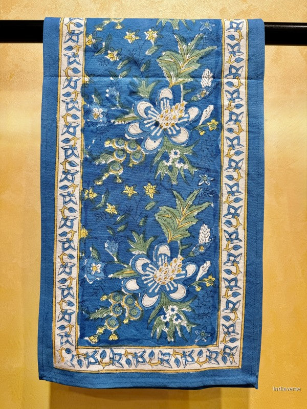 A square blue table runner featuring large white and blue flowers with green leaves and yellow accents, framed by an ornate patterned border on a tan background.