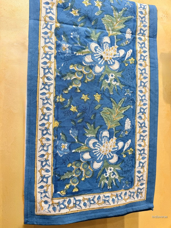 A square blue embroidered textile, likely a quilt or tapestry, featuring large white and blue flowers with green leaves and yellow accents, framed by an ornate patterned border on a tan background.