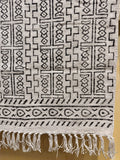 A close-up of a woven textile with a black geometric tribal pattern on a light beige or off-white background, finished with a fringed hem.