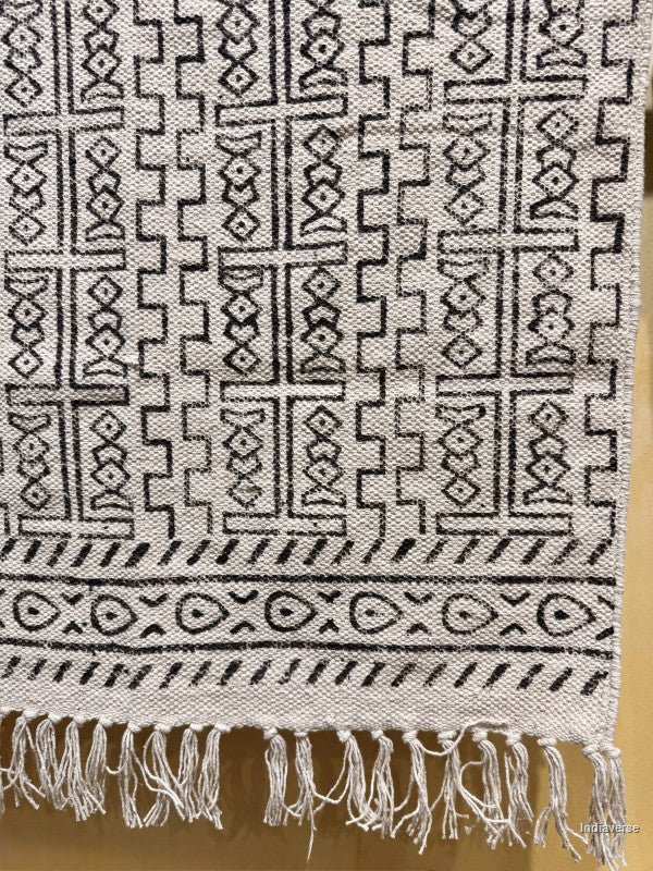 A close-up of a woven textile with a black geometric tribal pattern on a light beige or off-white background, finished with a fringed hem.