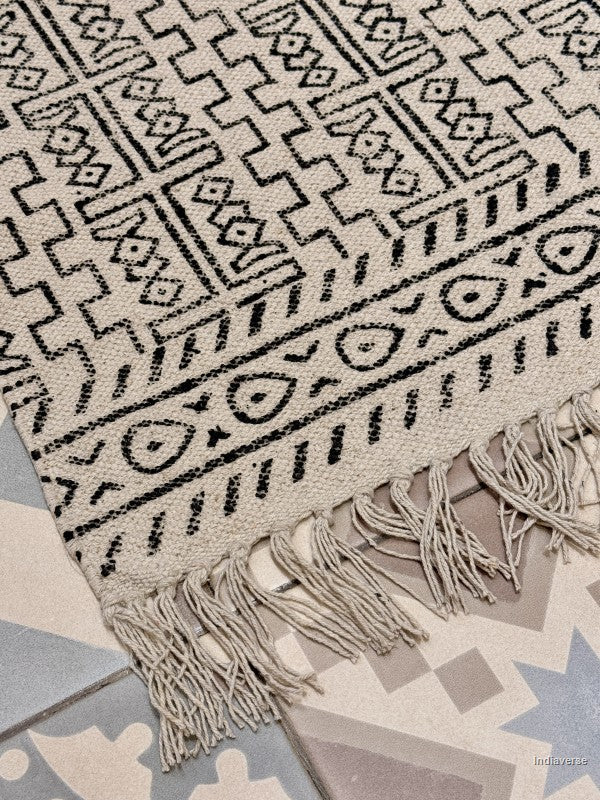 A close-up of a woven rug with a black geometric tribal pattern on a cream background, featuring fringed edges and shown laid over patterned floor tiles.