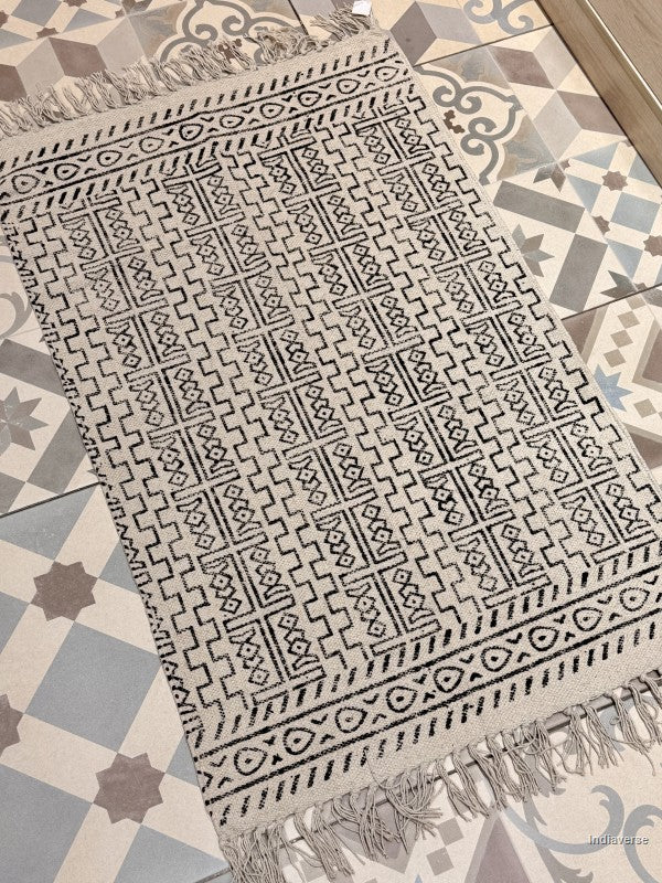 A rectangular woven rug with fringe, featuring a black geometric pattern on a light beige or off-white base, laid out on a patterned tile floor.