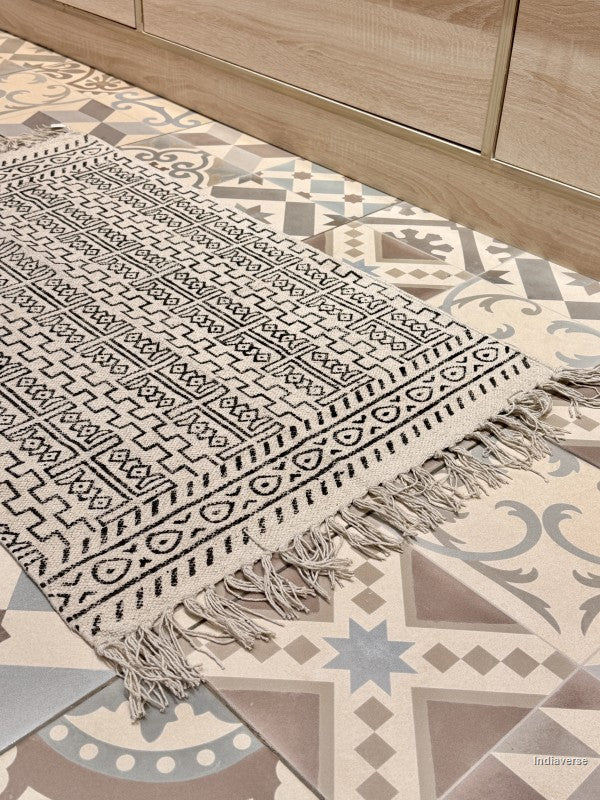 A rectangular woven rug with black geometric patterns and fringe tassels, laid out on a patterned tile floor next to a light wood cabinet.