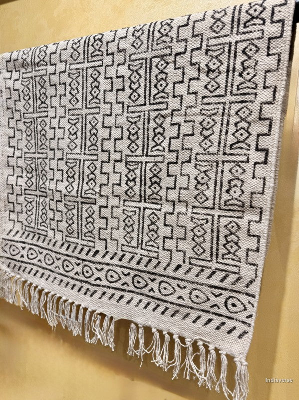 A woven textile with a black geometric tribal pattern on a light beige or off-white base, featuring a fringed hem, shown hanging against a warm beige background.