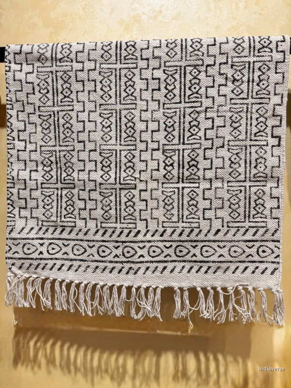 A woven textile with a black geometric tribal pattern on a light beige or off-white background, featuring a fringed hem, shown hanging against a warm beige wall.