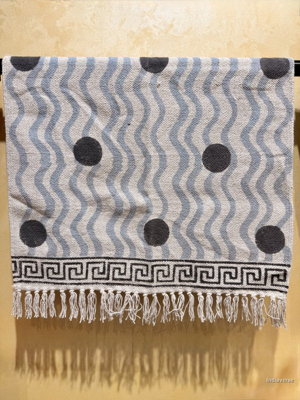 A woven textile throw blanket with a gray and white wavy pattern, black polka dots, and a black Greek key border with white fringe tassels, hanging against a beige wall.