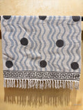 A woven textile throw blanket with a gray and white wavy pattern, black polka dots, and a black Greek key border with white fringe tassels, hanging against a beige wall.