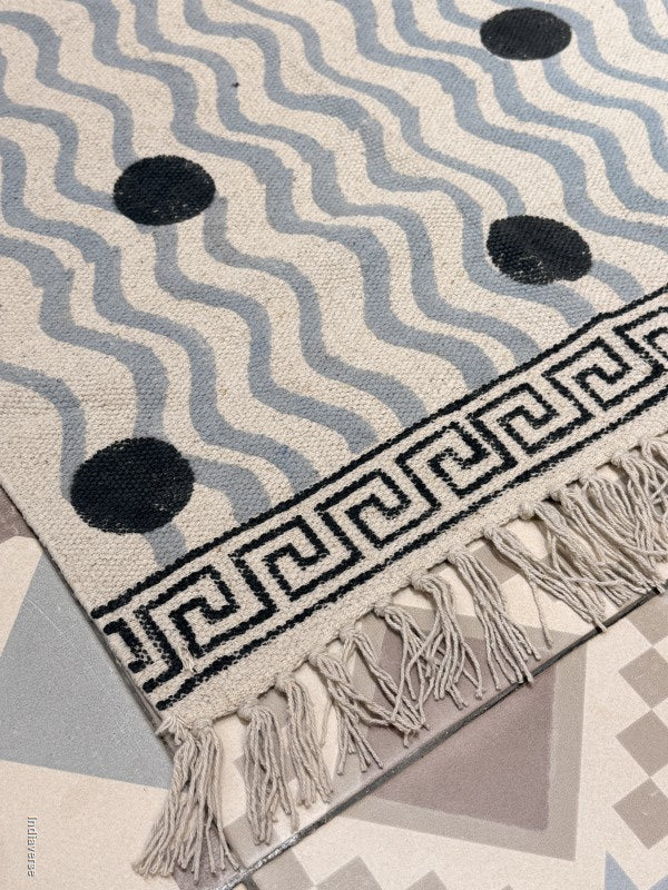 A close-up of a woven rug with a cream base, light blue wavy stripes, black circular motifs, a black Greek key border, and off-white fringe tassels, laid over a patterned floor.