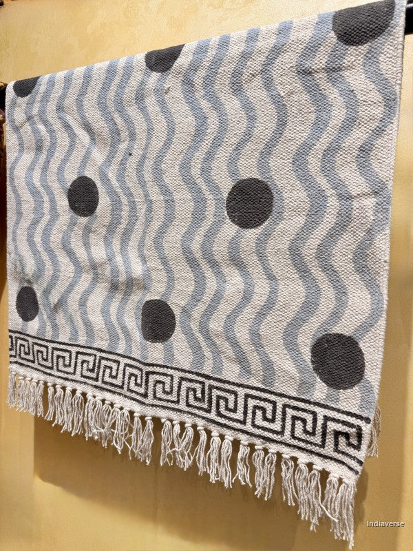 A woven textile throw or blanket with a gray and white wavy stripe pattern, black polka dots, a black Greek key border, and white fringe tassels, hanging against a warm beige wall.