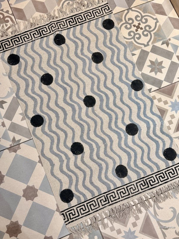 A rectangular area rug with a light beige base, featuring a pattern of wavy blue-gray stripes, black polka dots, and black Greek key borders with fringe on the ends, laid out on a patterned tile floor.